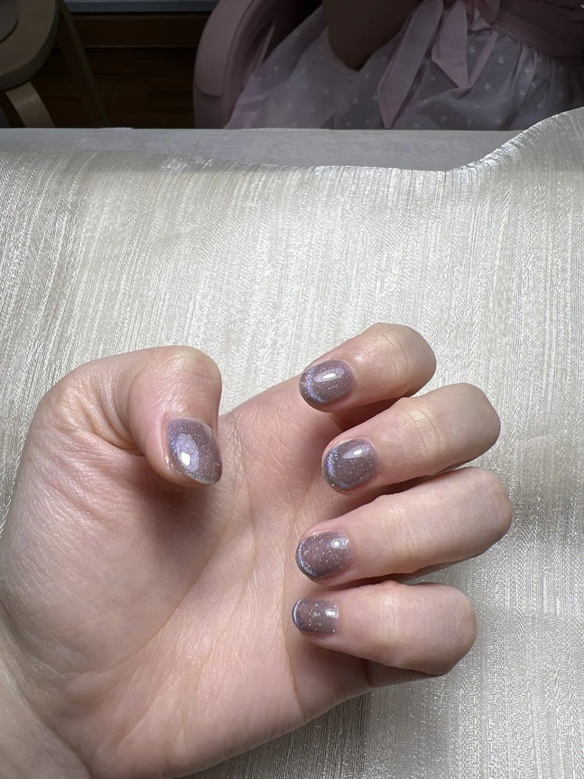 Close-up of a hand with nails painted in a glittery, lavender color resting on a textured, cream-colored surface. In the background, part of a woman in a pink hospital gown is visible.