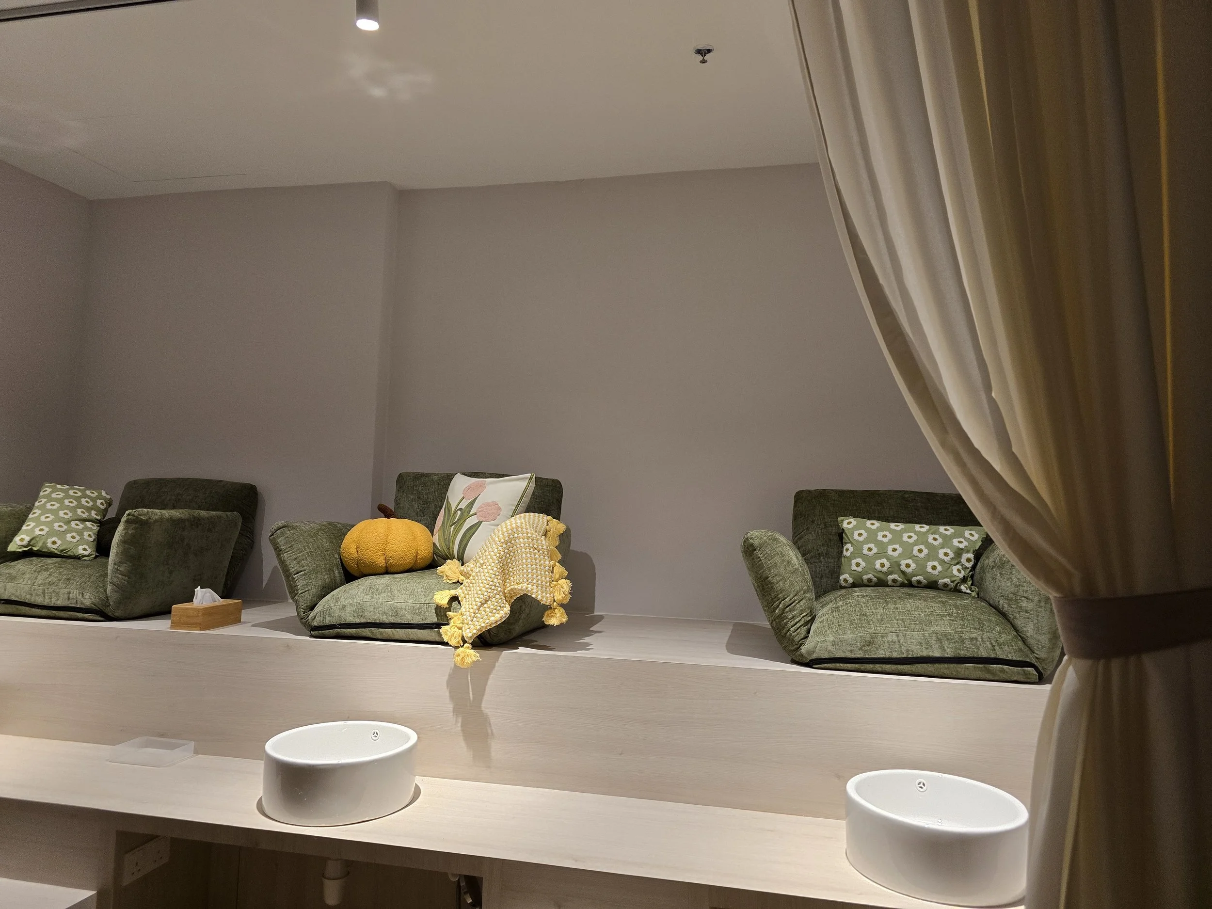 Interior view of a room with a gray wall, green cushions on the sofa, and decorative pillows with floral patterns. The room has a white table with two white round objects on it, possibly humidifiers or air purifiers. Part of a curtain is visible on the right side of the image.