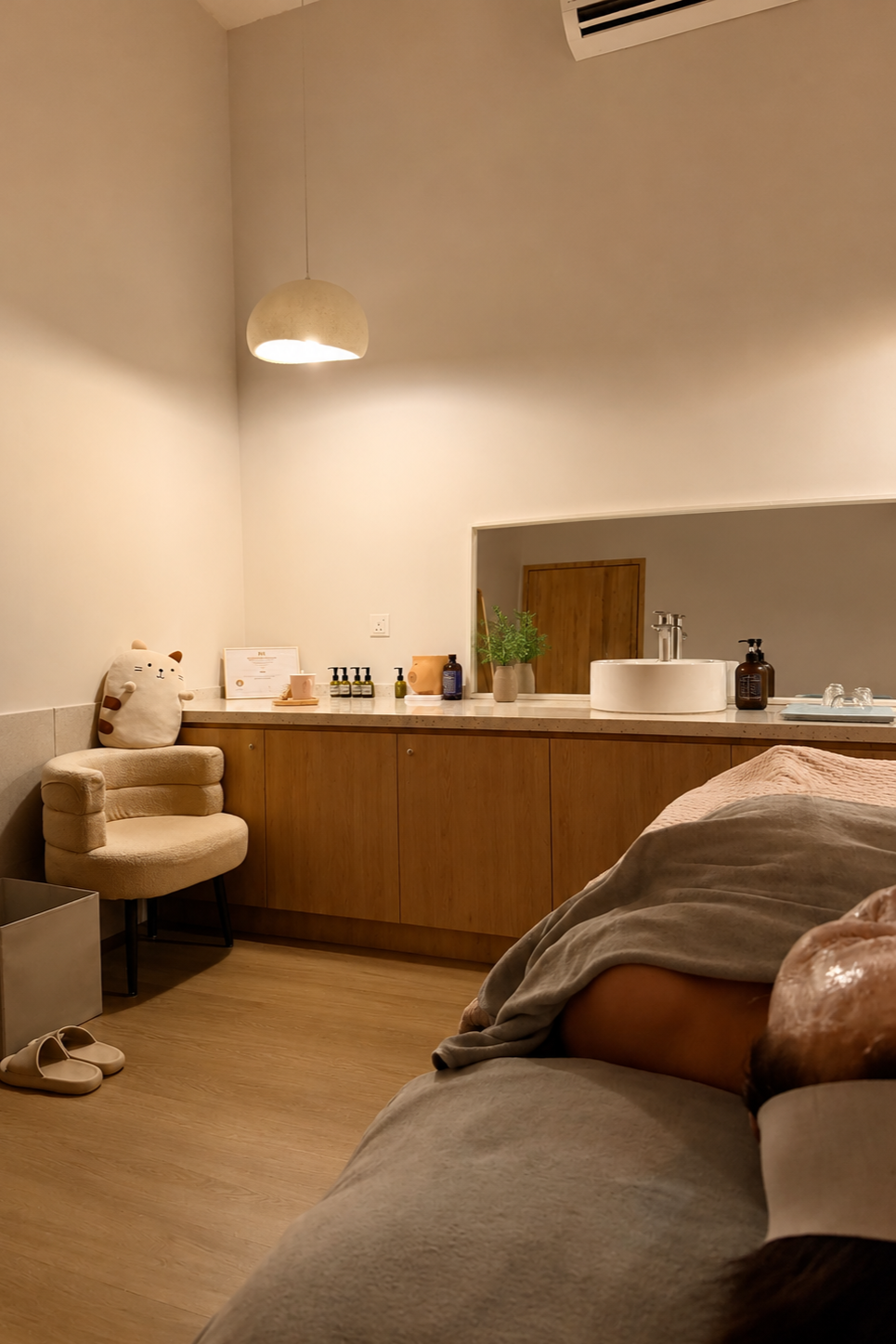 Interior of a cozy, softly lit spa or treatment room with a wooden cabinet, a plush chair with a cat-shaped pillow, bottles of products, a potted plant, and a lying down person in a relaxing setting in Getsumei Beauty.