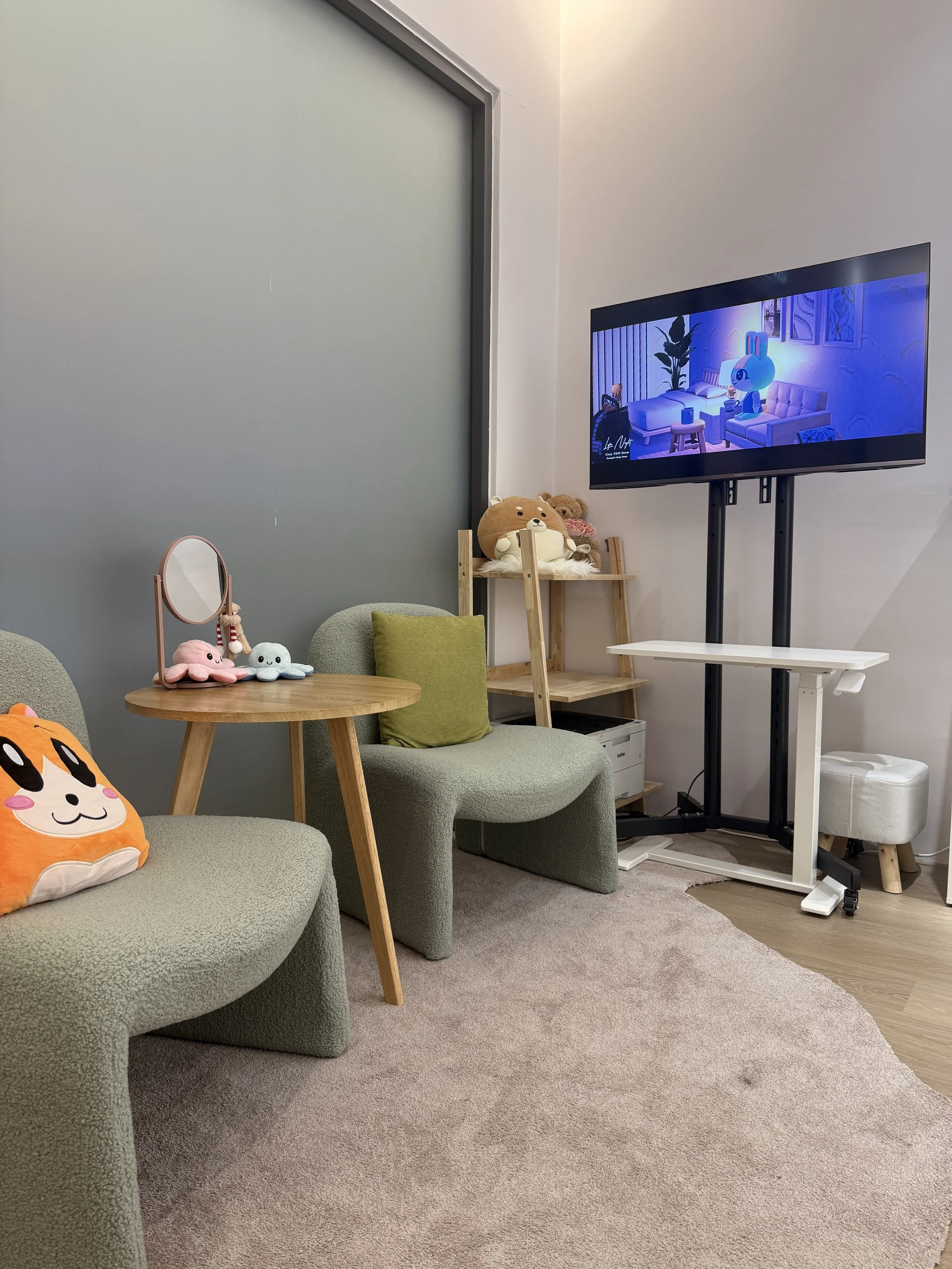 A cozy room with a green textured sofa and a circular wooden side table holding plush toys, including a pink starfish, a white cloud, and an orange animal. There is a small mirror on the table. A green pillow is on the sofa. In the background, a wooden shelf holds teddy bears. A large flat-screen TV is mounted on a stand