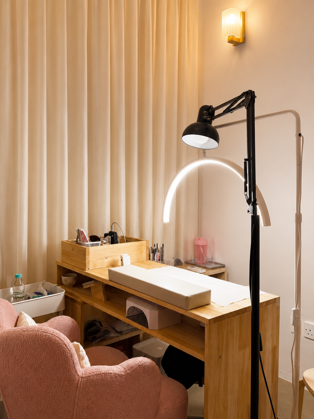 A manicure station in Getsumei Beauty with a wooden table, pink armchair, and a lamp with a black and a white adjustable arm, set against beige curtains and a wall-mounted light.