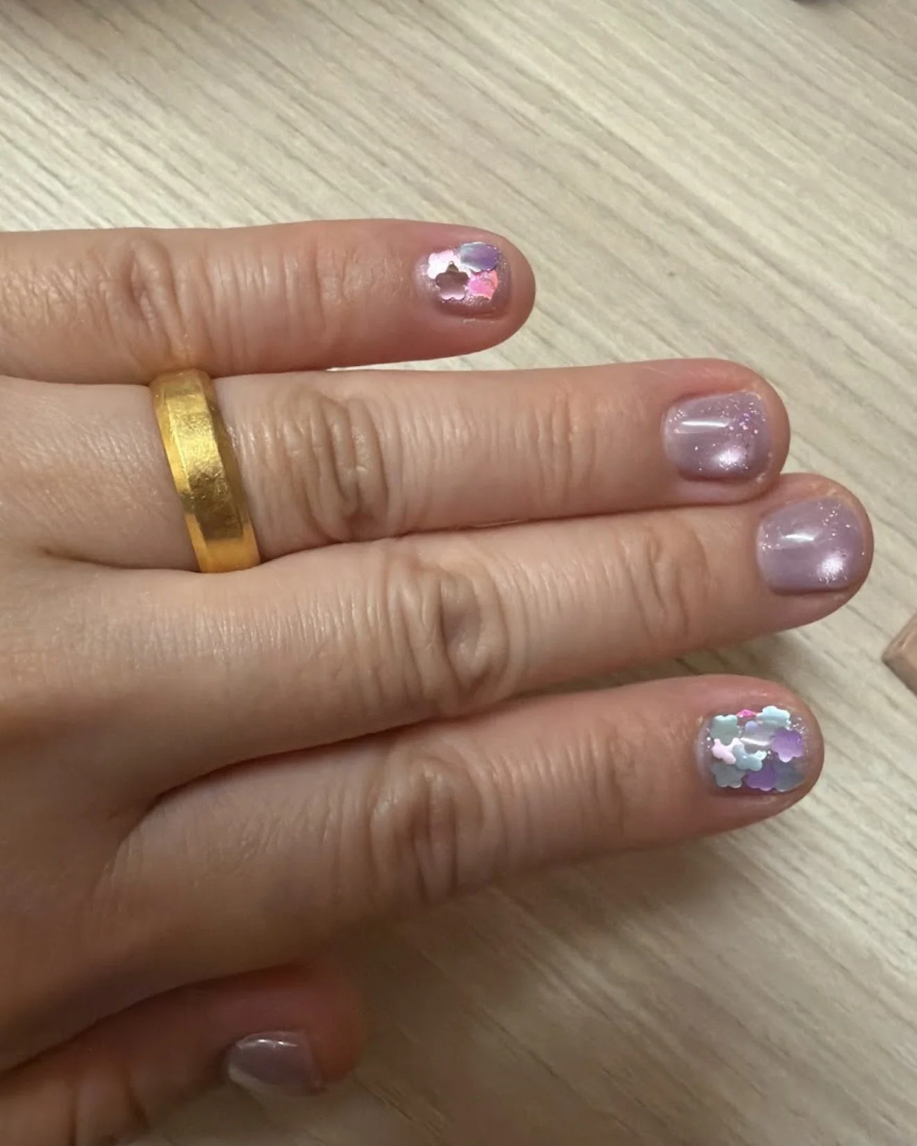 Close-up of multiple fingers with colorful glittery nail polish and a gold ring on the middle finger, resting on a light wooden surface.