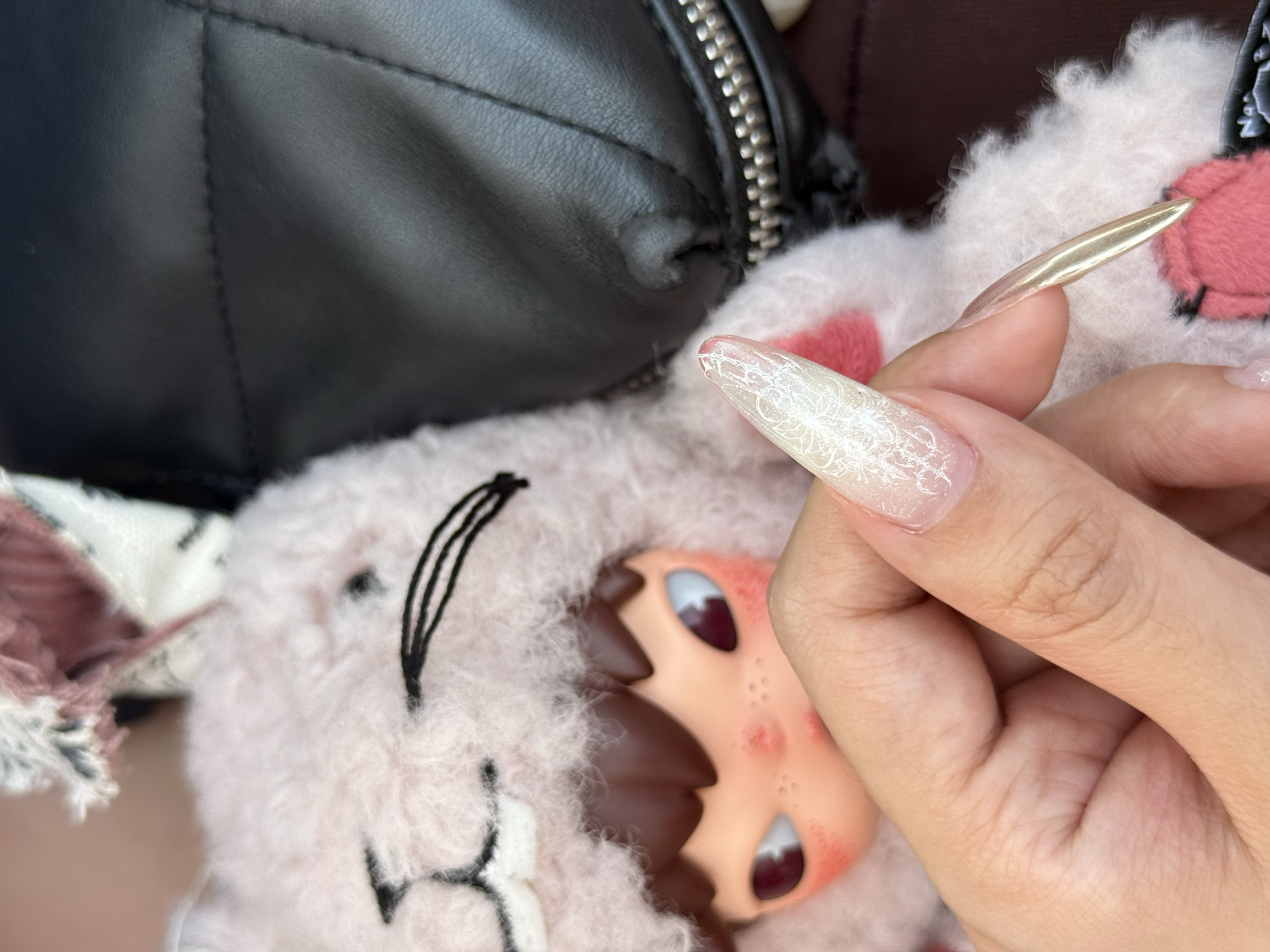 Close-up of a hand with long, decorative, metallic-colored nails touching a stuffed animal with pink and white fur, near a black leather bag with a silver zipper.