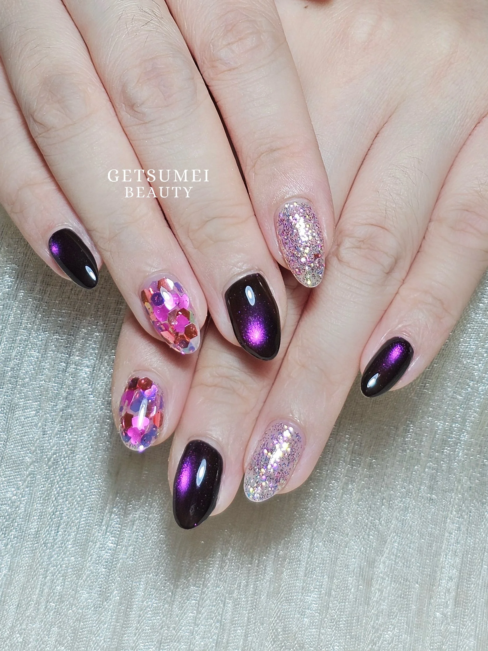 Close-up of a woman's hand with manicured nails on a silver textured fabric background. The nails feature designs including dark purple, glitter, and rhinestones. Text overlay reads 'GETSUMEI BEAUTY'.