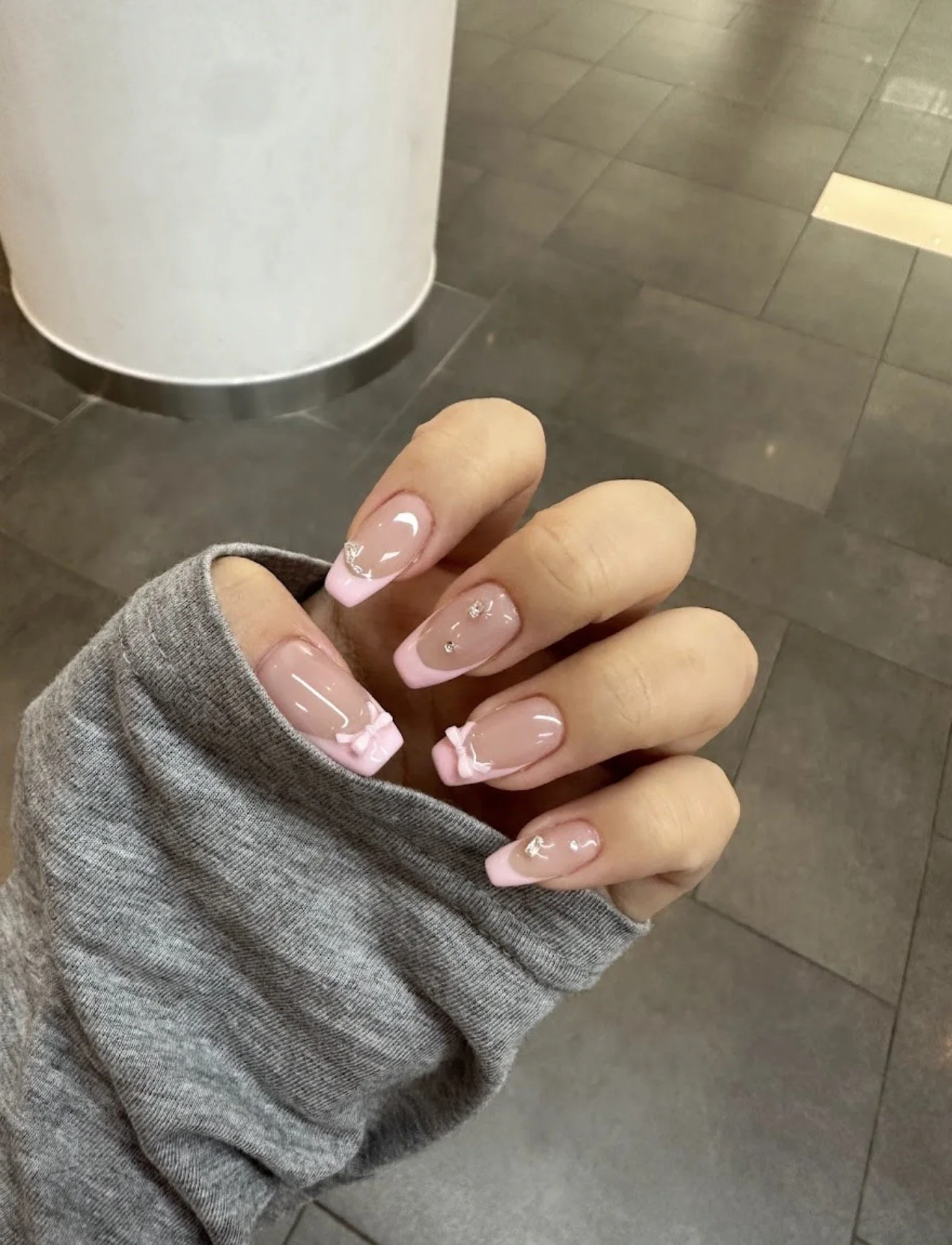 Close-up of a woman's hand with manicured nails featuring pink tips and small decorative elements, wearing a gray sweater.