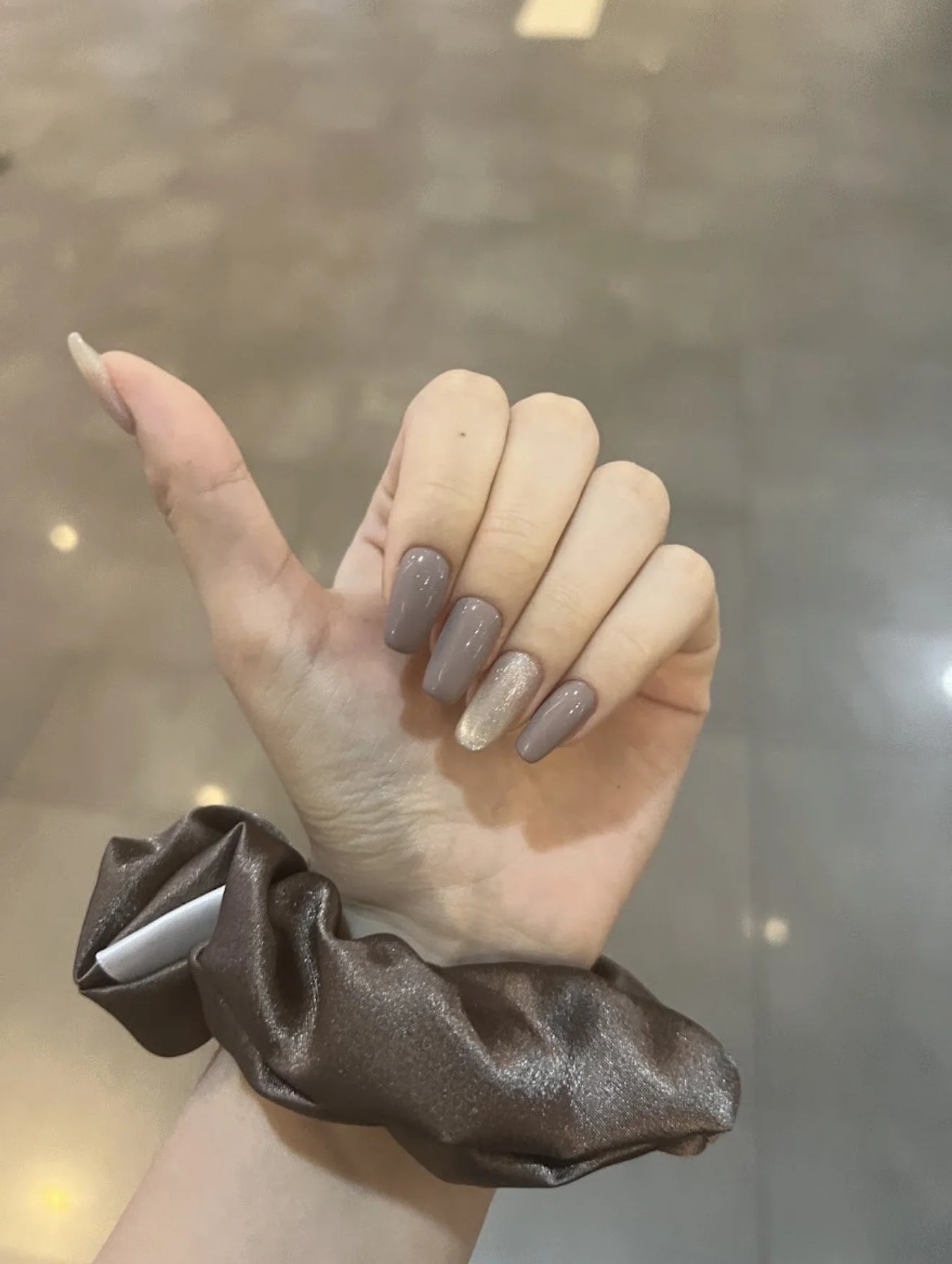 A person's hand showing off manicure with nude and glittery nail polish, wearing a brown scrunchie with a shiny finish.