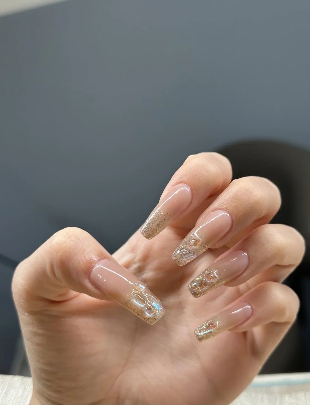 Close-up of a hand with long, decorated nails featuring a clear gloss, gold glitter, and holographic accents.