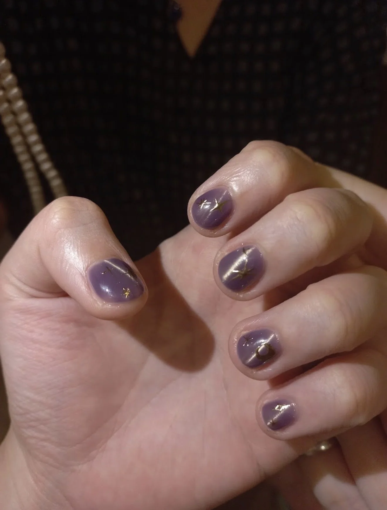 Close-up of a person's hand showing short nails painted with translucent purple nail polish with small gold star and crescent moon decorations.