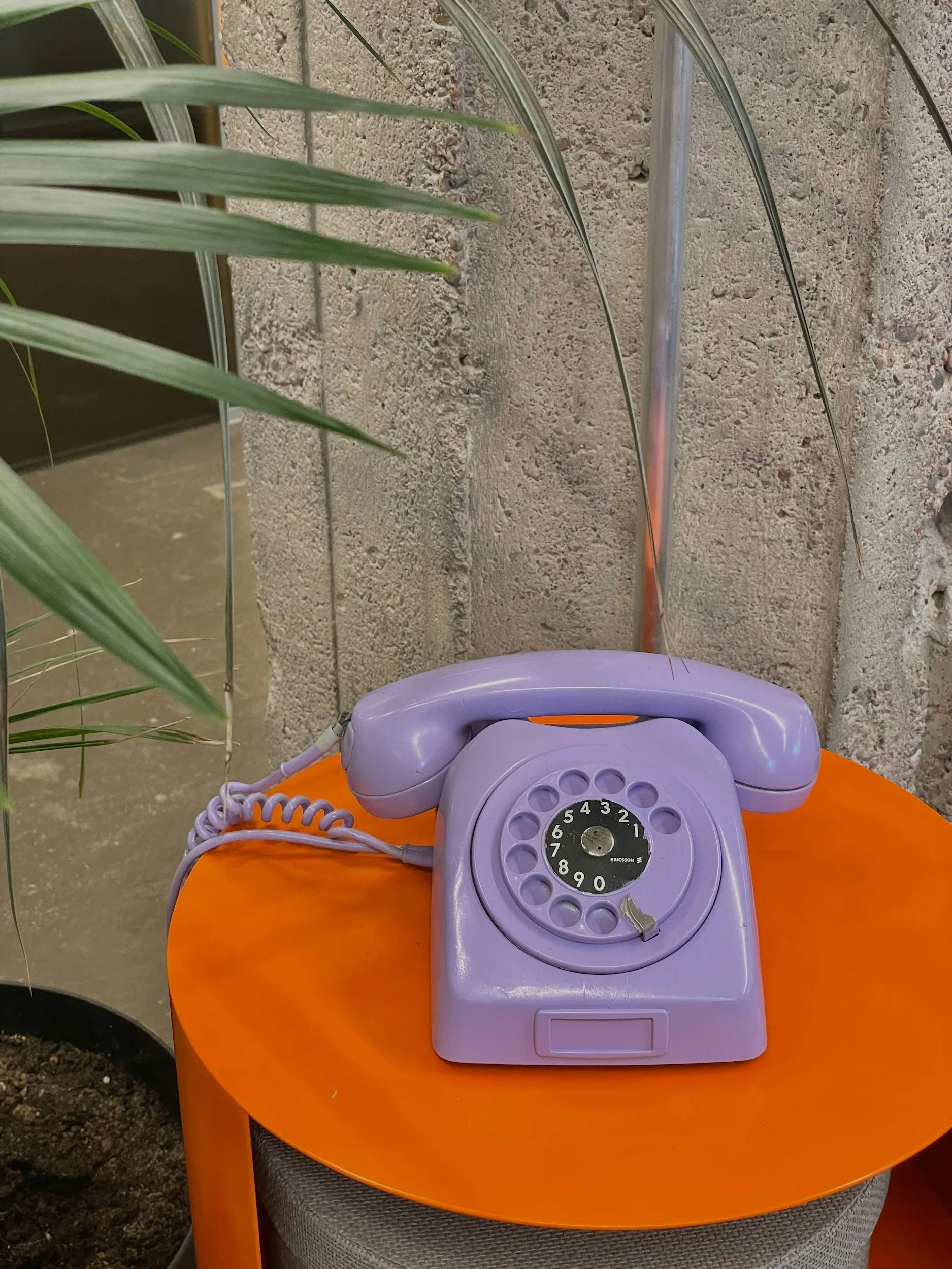 A purple rotary dial telephone resting on an orange round table, with green plant leaves partially visible in the background.