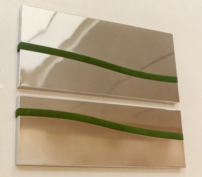 Two rectangular wall art pieces with metallic surfaces and a curved green line across each, hung vertically on a white wall.