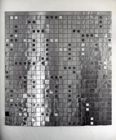 A mosaic-style artwork composed of small square tiles in various shades of gray arranged in a pattern, with a central vertical light streak on a reflective, textured surface.