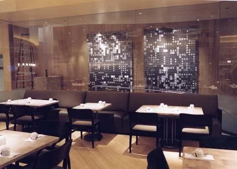 Empty restaurant dining area with tables set for guests, dark chairs, and abstract black and white artwork on the wooden wall in the background.