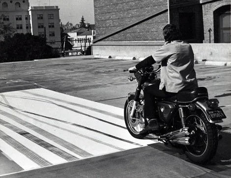 Man sitting on a motorcycle on a rooftop parking lot with buildings in the background.