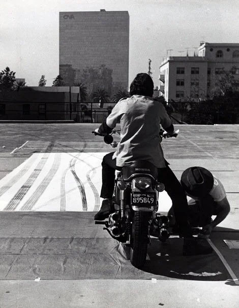 A person on a motorcycle drawing tire skid marks on a parking lot surface, with a second person crouching next to them. In the background, there are buildings and a large office tower.
