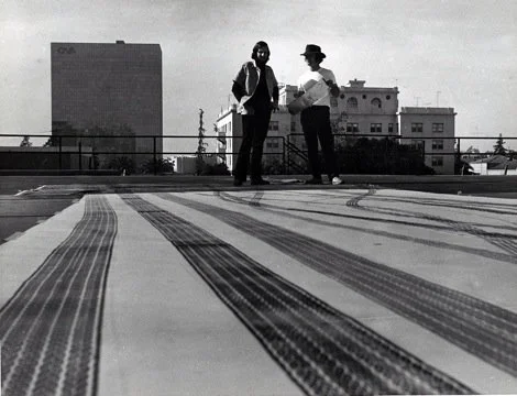 Two women standing on a rooftop with city buildings in the background, viewed from a low angle near a patterned rooftop surface.