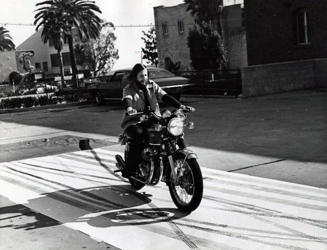 Person riding a motorcycle across a street in a residential area with trees and buildings in the background.
