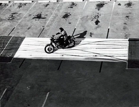 A person riding a motorcycle on a striped crosswalk in a parking lot.