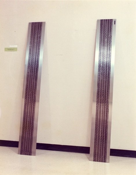 Two tall, narrow, silver-colored sculptures with intricate dark patterning, standing on a white floor against a plain white wall in an art gallery.