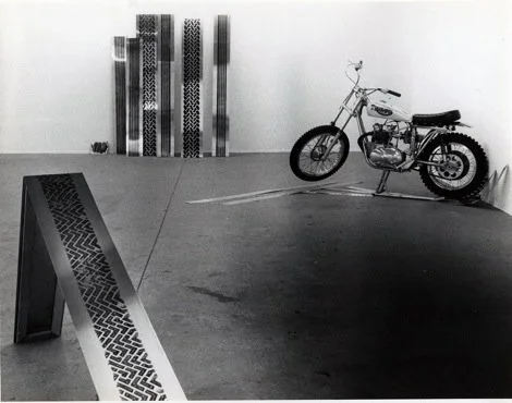 An art gallery with a bicycle on display, abstract patterned artworks on the wall, and a patterned stand in the foreground.