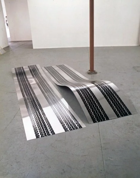 A piece of paper with black and white striped patterns is crumpled on a gray tile floor in an empty room with a white wall and a orange pole.