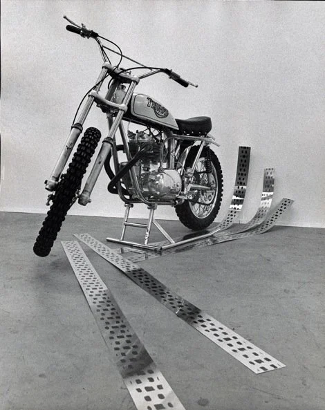 A vintage snowmobile with metal skis and large tires, positioned indoors against a plain wall.