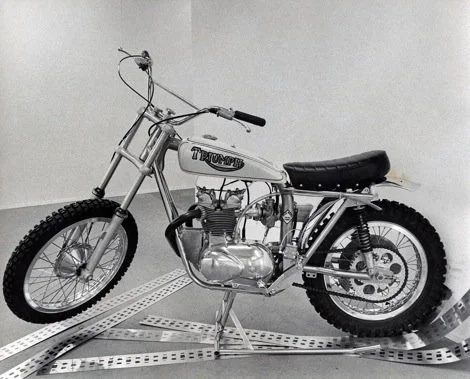 Vintage Triumph motorcycle with a silver frame, black seat, and knobby tires, positioned on a metal grid surface in front of a plain white wall.