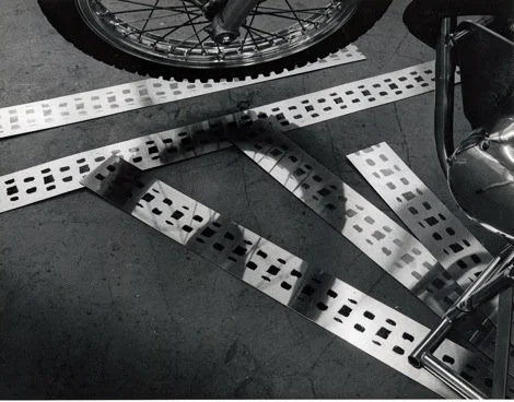 A white metal parking barrier with multiple rectangular holes, lying on a dark surface next to a motorcycle wheel and chrome handlebars.