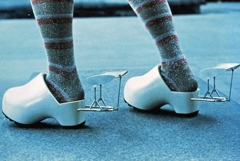 Close-up of a person wearing sparkly striped socks and white clown shoes with metal clickers attached, standing on a blue surface.