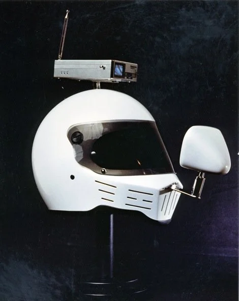 A white motorcycle helmet with added electronic equipment, including a small screen, antenna, and a side-mounted mirror, against a dark background.