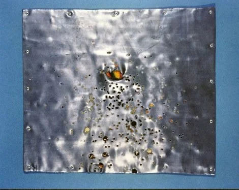 A metal sheet with water droplets and a falling water droplet creates a splash pattern, mounted on a blue wall.