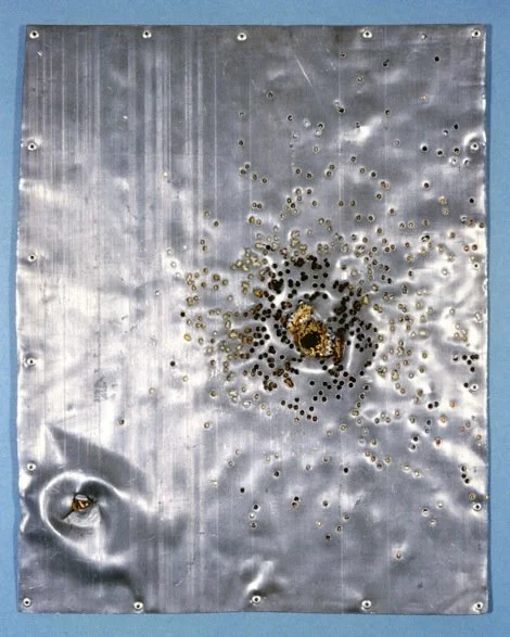 Metal sheet with burn marks and multiple small holes, some showing signs of melting or deformation, against a blue background.