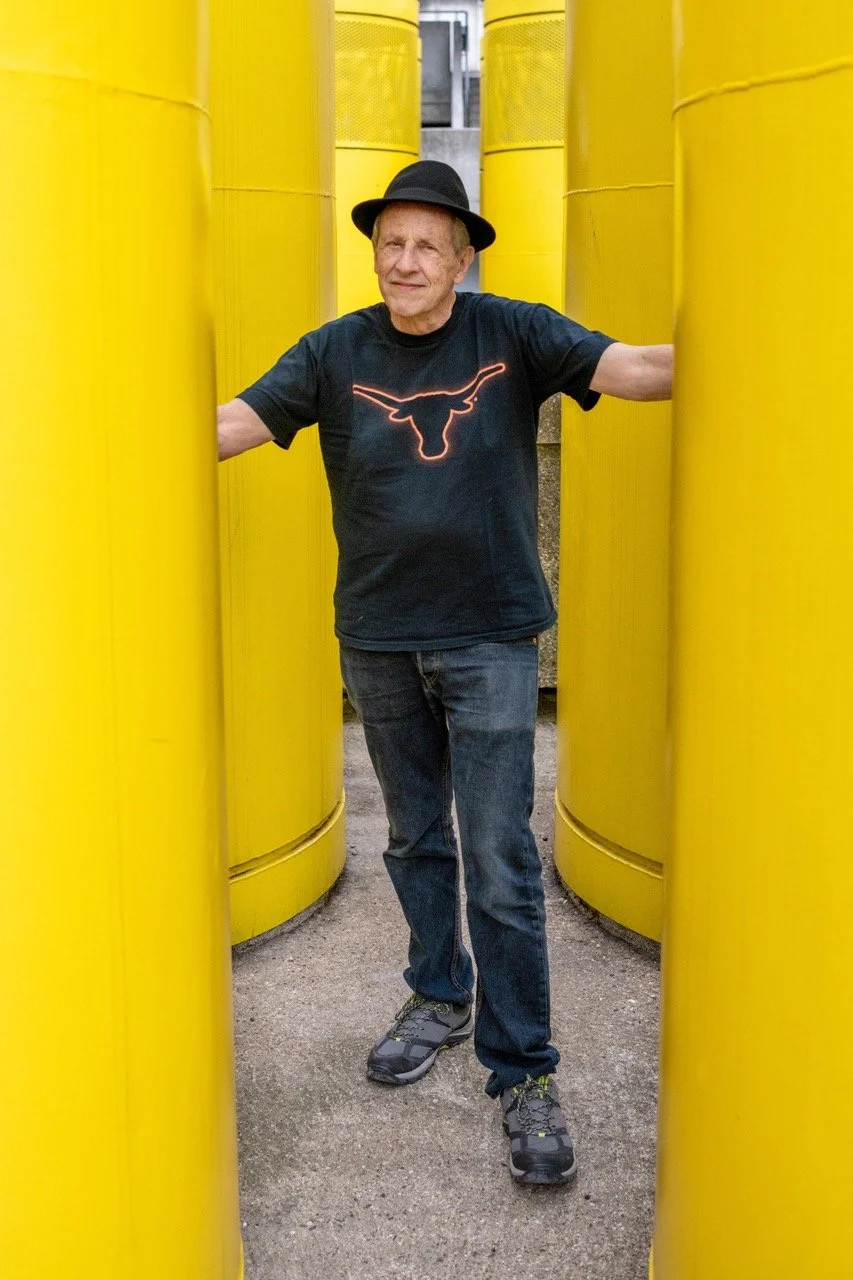 A man wearing a black hat, black t-shirt with a neon Texas Longhorns logo, dark jeans, and sneakers, standing between large yellow industrial tanks.