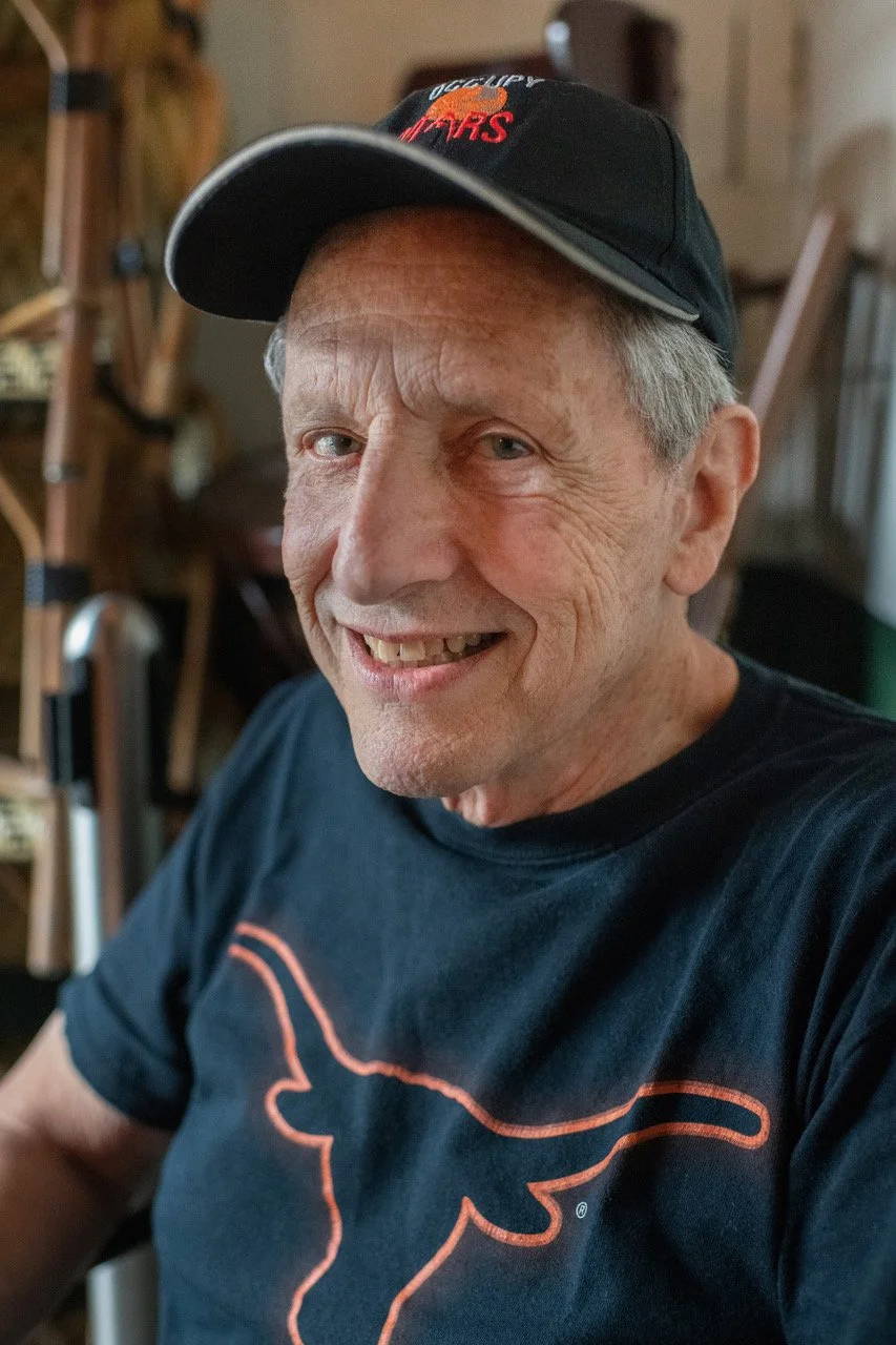 An elderly man smiling, wearing a black cap and a black T-shirt with a longhorn outline on it, sitting indoors with blurred background.