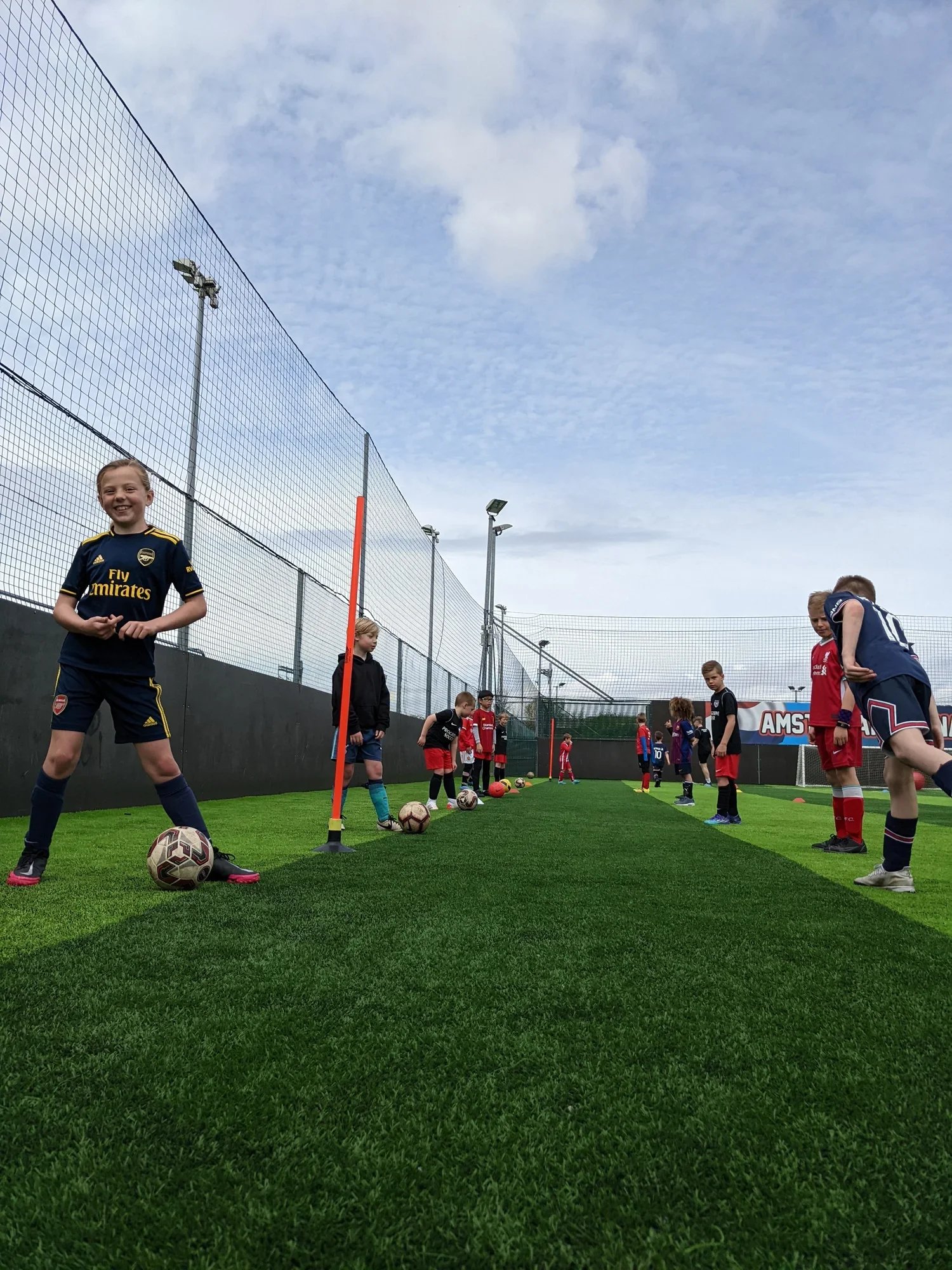 Children practicing football on an outdoor field with a group of kids standing behind a training line with football balls, wearing sports kits at a Progressive Soccer Half-term Camp