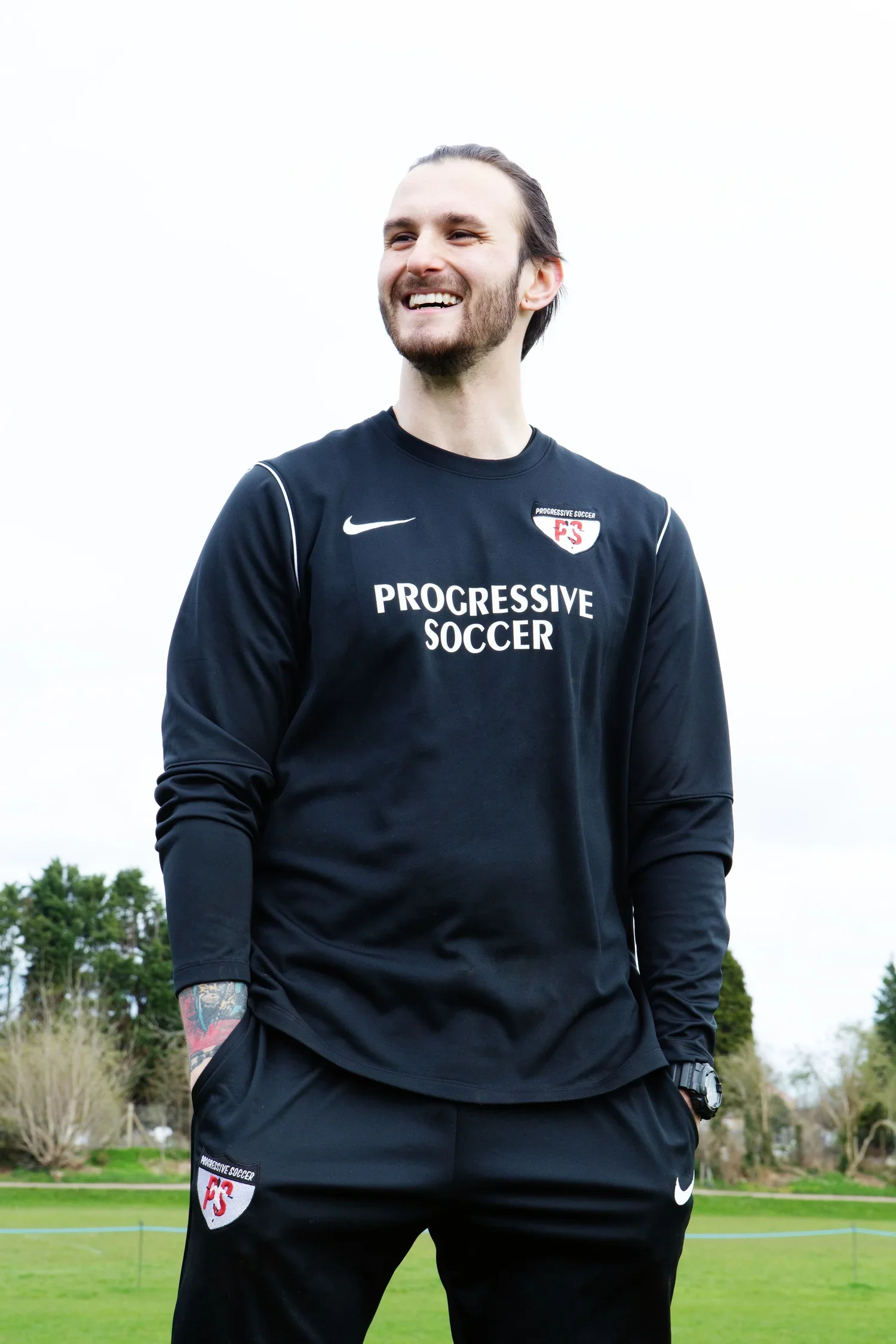 A coach with long hair, beard, black sportswear with 'Progressive Soccer' logo, standing outdoors on a grassy field, smiling, with trees in the background.