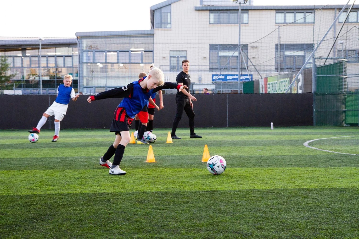 Young boys practicing football drills on an outdoor field with yellow cones, under the supervision of a coach, with a building and netting in the background.