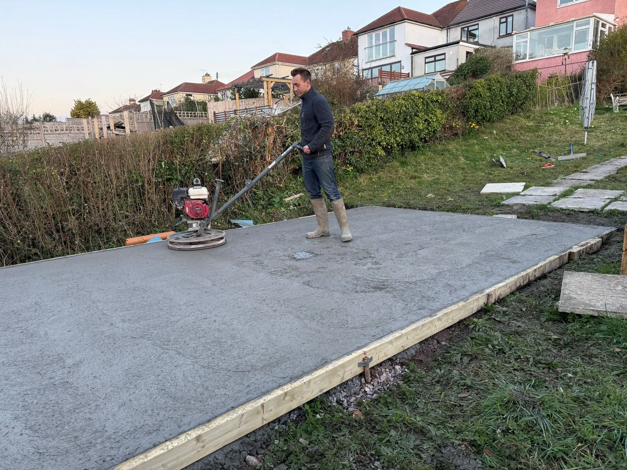 A person wearing boots using a concrete power trowel on freshly poured concrete in a backyard with houses and bushes in the background.