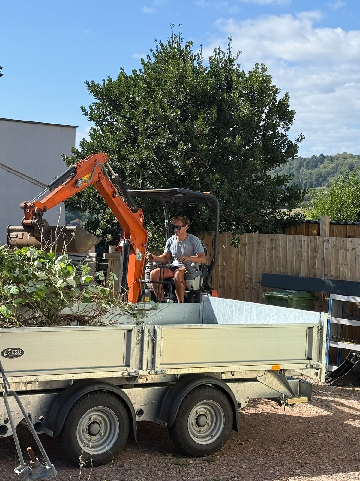 A person operating a small orange excavator on a trailer filled with plants and debris in a backyard with a wooden fence and a large tree in the background.