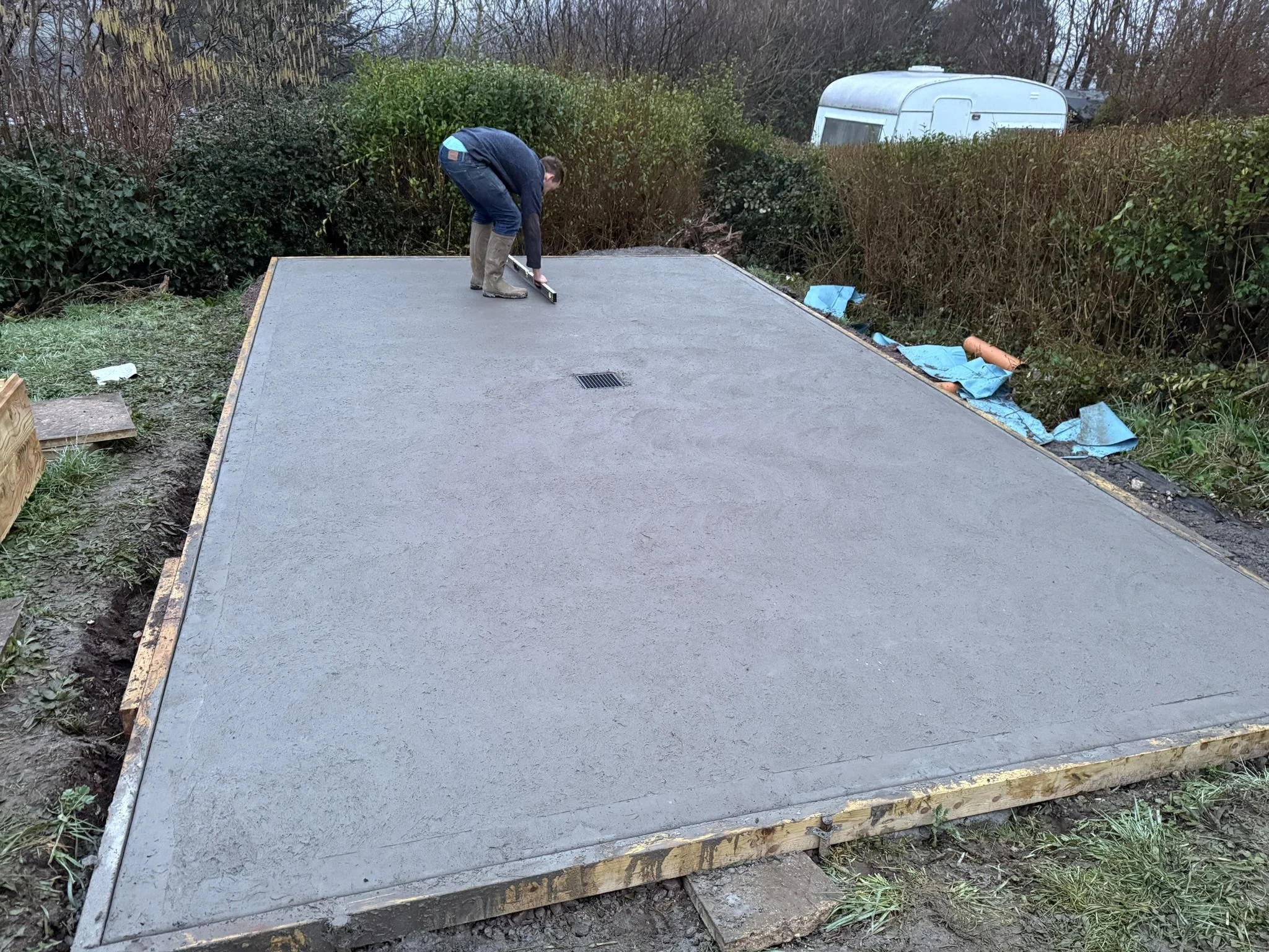 A man is smoothing fresh concrete on a wooden form with a trowel, preparing a concrete slab outdoors surrounded by bushes and trees, with a white trailer visible in the background.