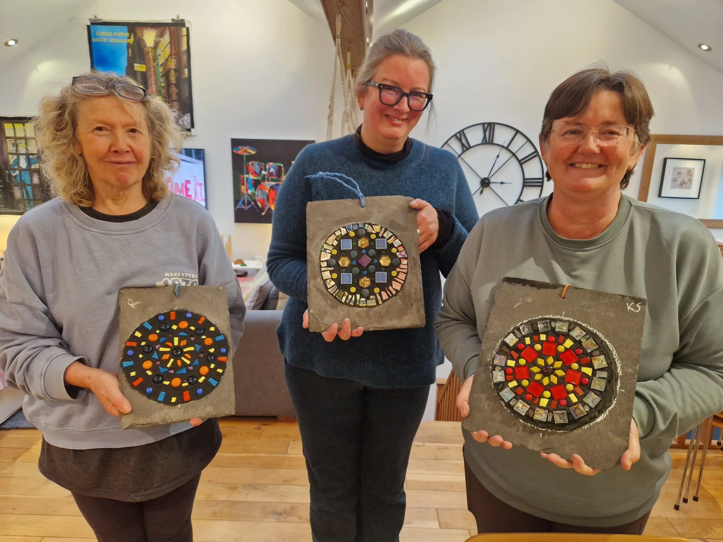 Three women standing indoors, each holding a piece of art with circular mosaic designs on stone slabs, smiling at the camera. The background features a large clock on the wall and artwork pieces.