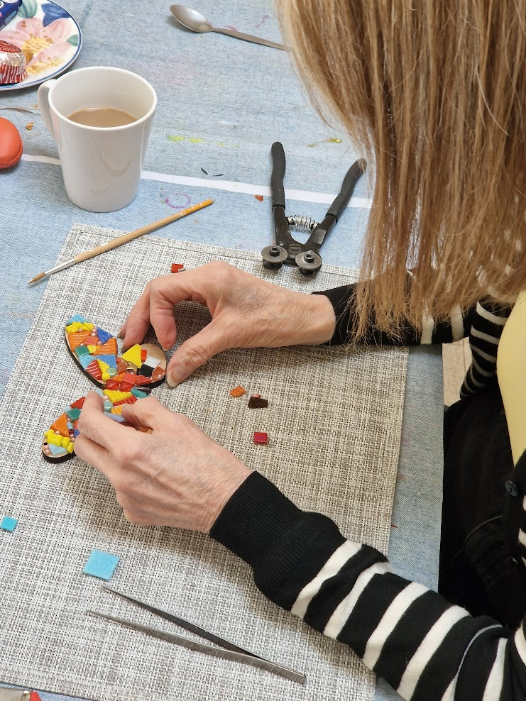 Person working on mosaic butterfly art project with small colorful tiles, tools, and a cup of coffee on the table.