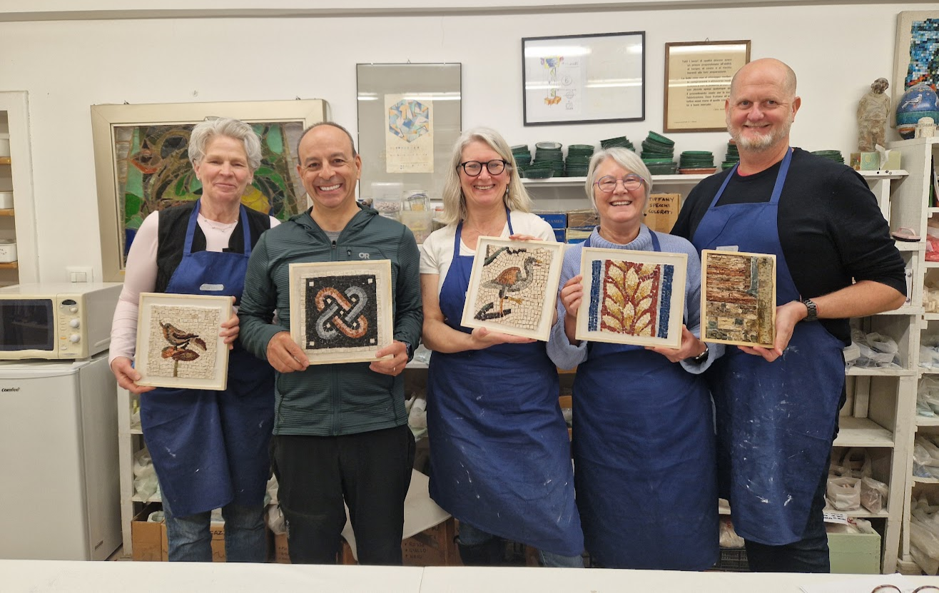Jacky and her peers at the Luciana Notturni Art School, each holding a framed mosaic artwork. They are wearing aprons in a studio, with shelves, framed pictures, and art supplies in the background.