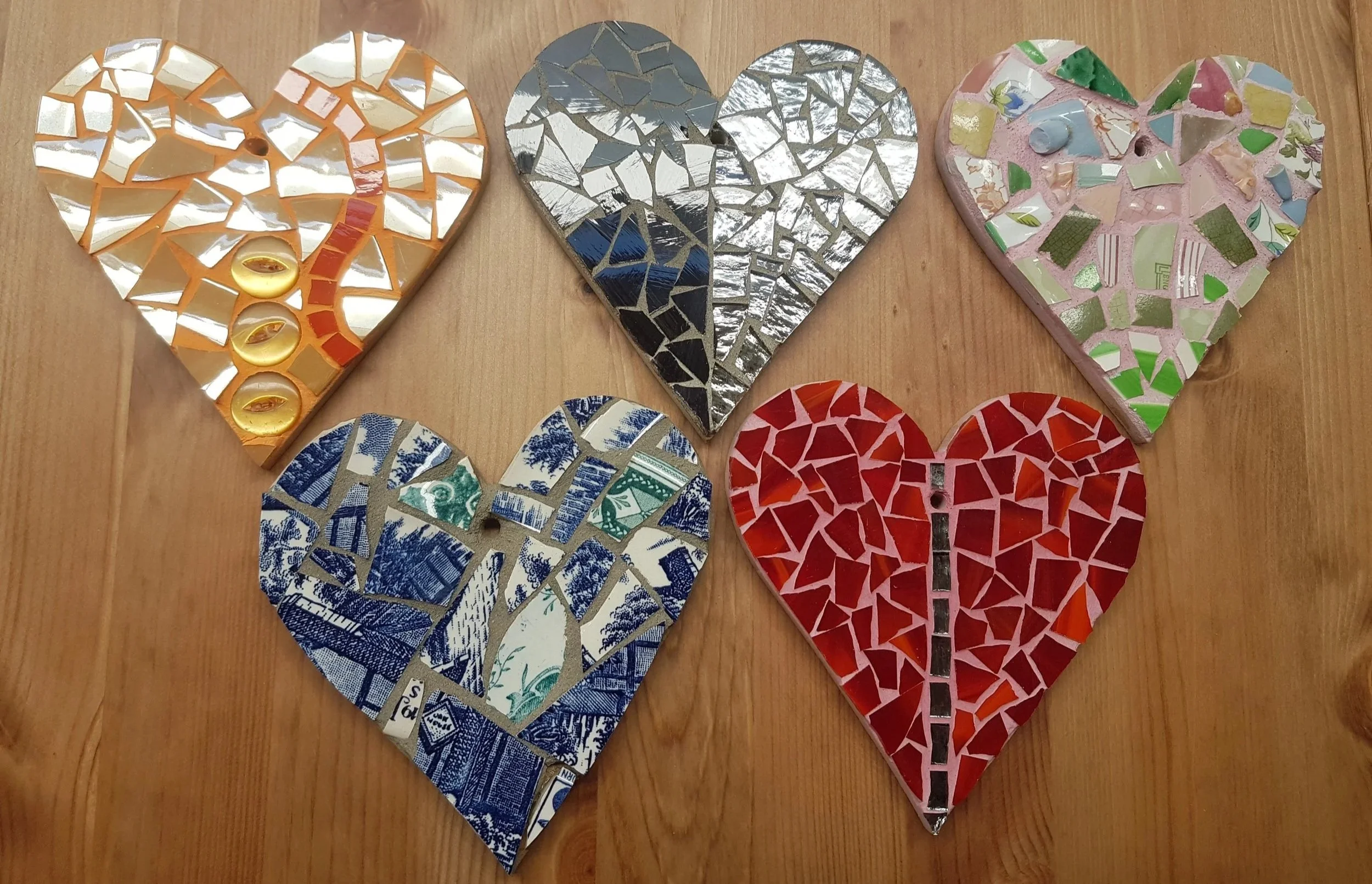 Six heart-shaped mosaics arranged in a row on a wooden surface. Each heart is made of broken mirror or ceramic pieces with different colors and patterns.