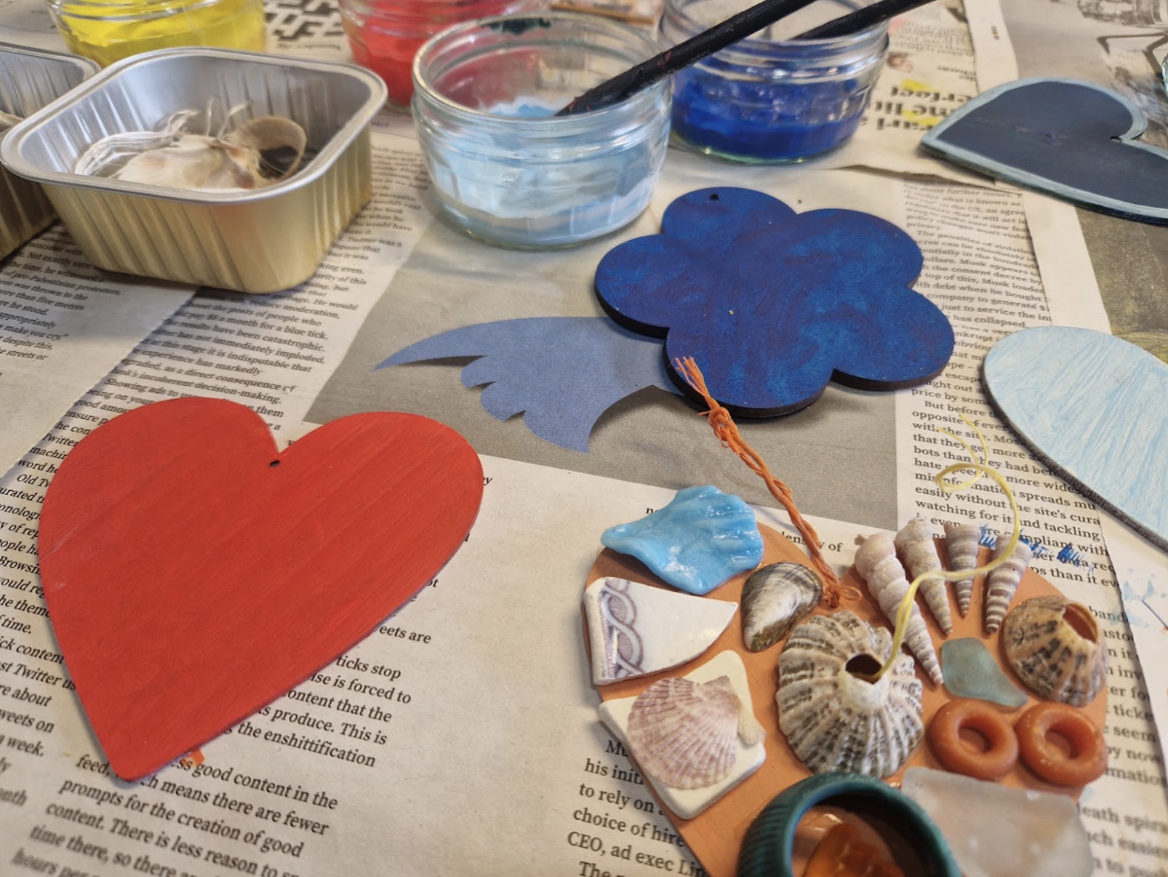 Craft table with painted paper hearts, seashells, shells, shells, shells, a starfish, and paint jars with brushes, on top of newspaper.
