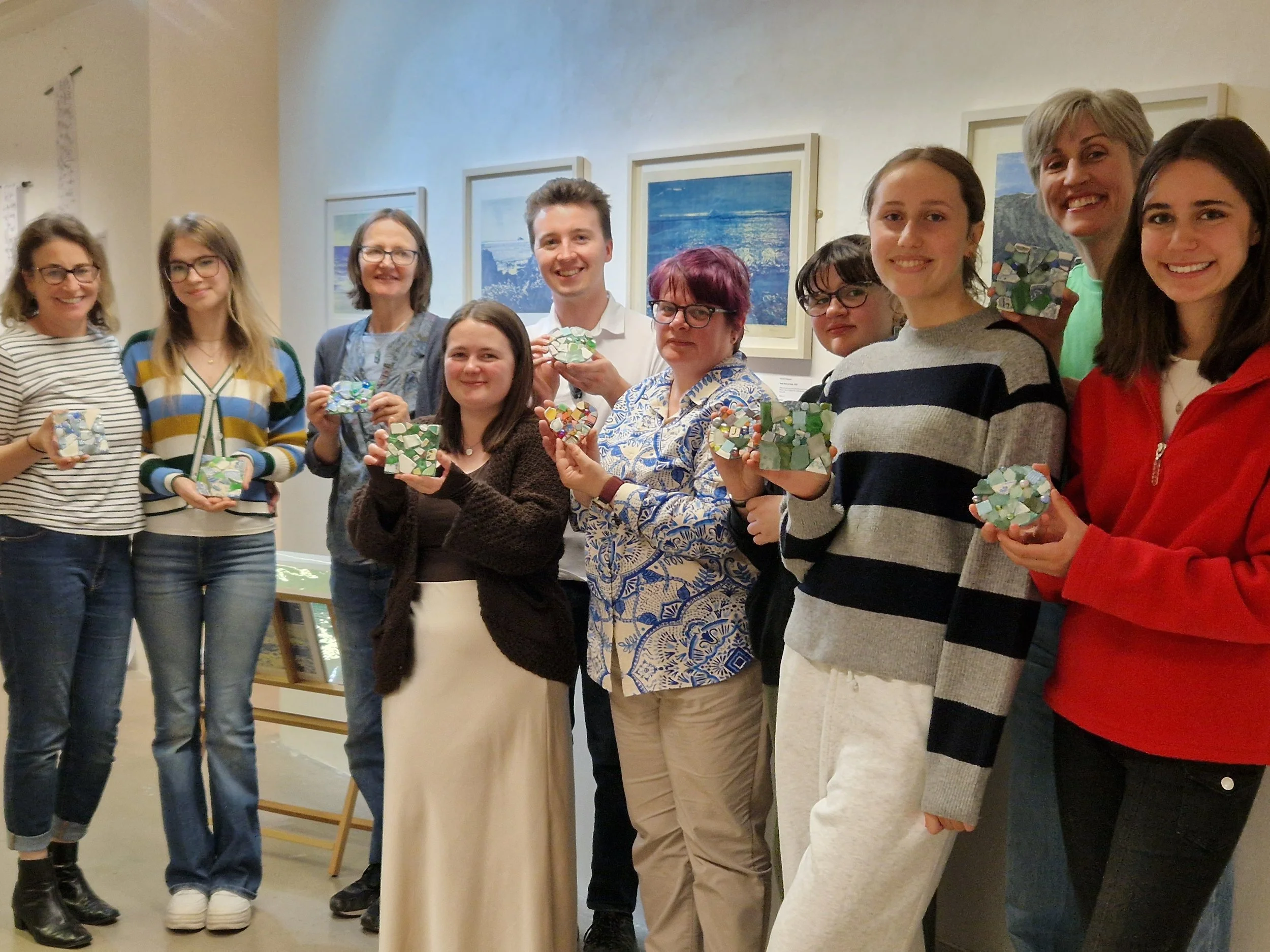 Group of ten people standing together indoors, holding mosaic-style artwork, smiling for the photo, with framed landscape pictures on the wall behind them.