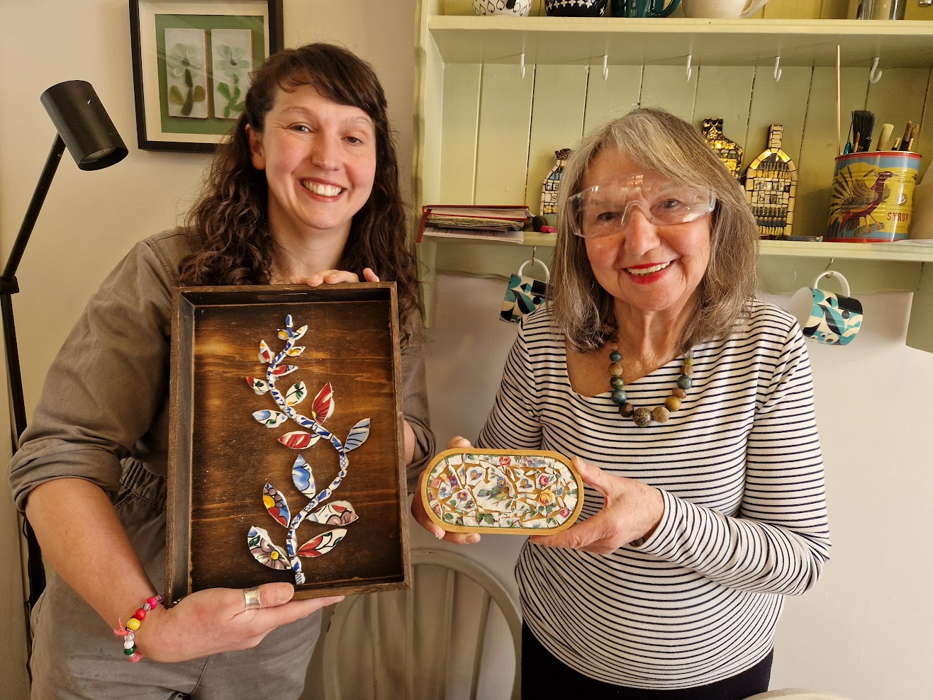 Two participants, holding decorative art pieces; the woman on the left holds a wooden framed art piece with a colourful floral design, and the woman on the right holds a small decorated box.