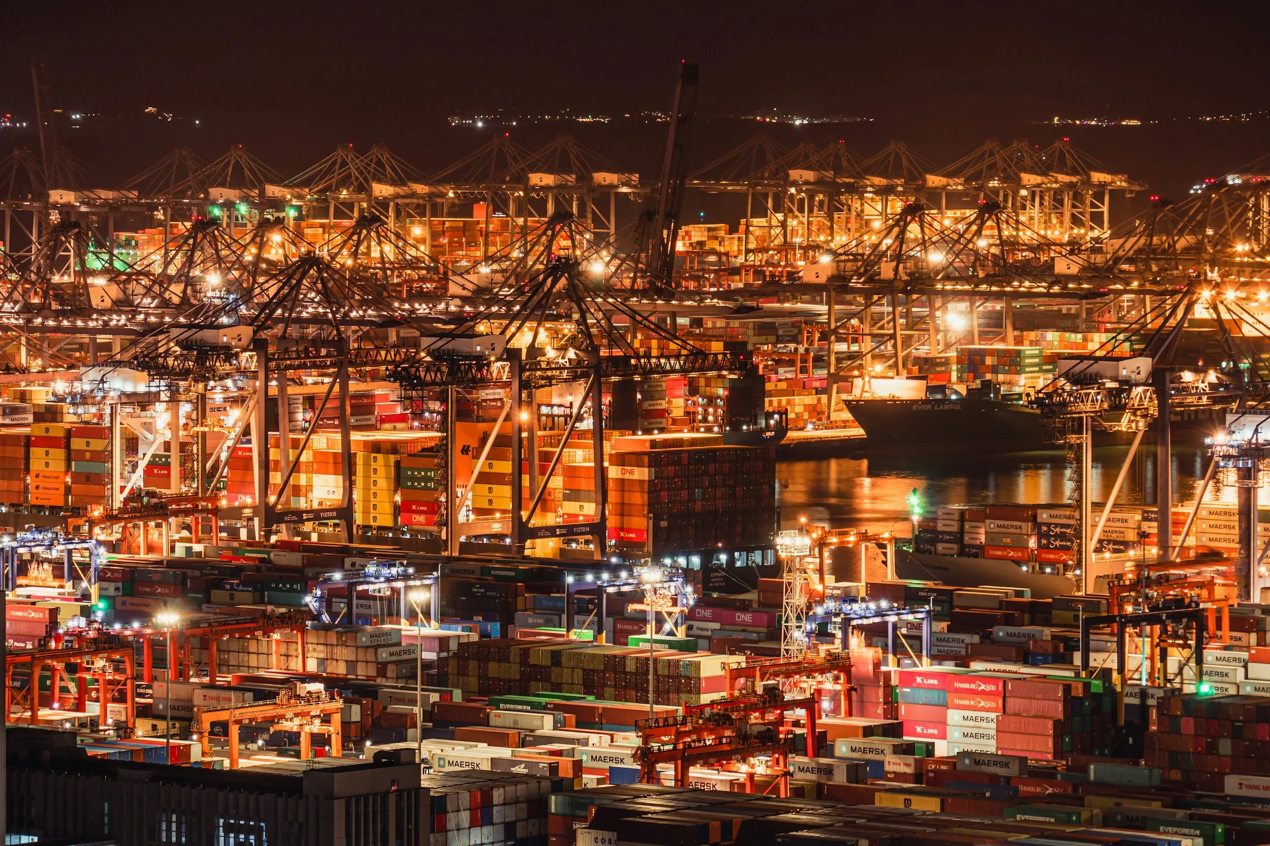 Night view of a busy shipping port with numerous stacked shipping containers, large cranes, and cargo ships illuminated by bright lights.