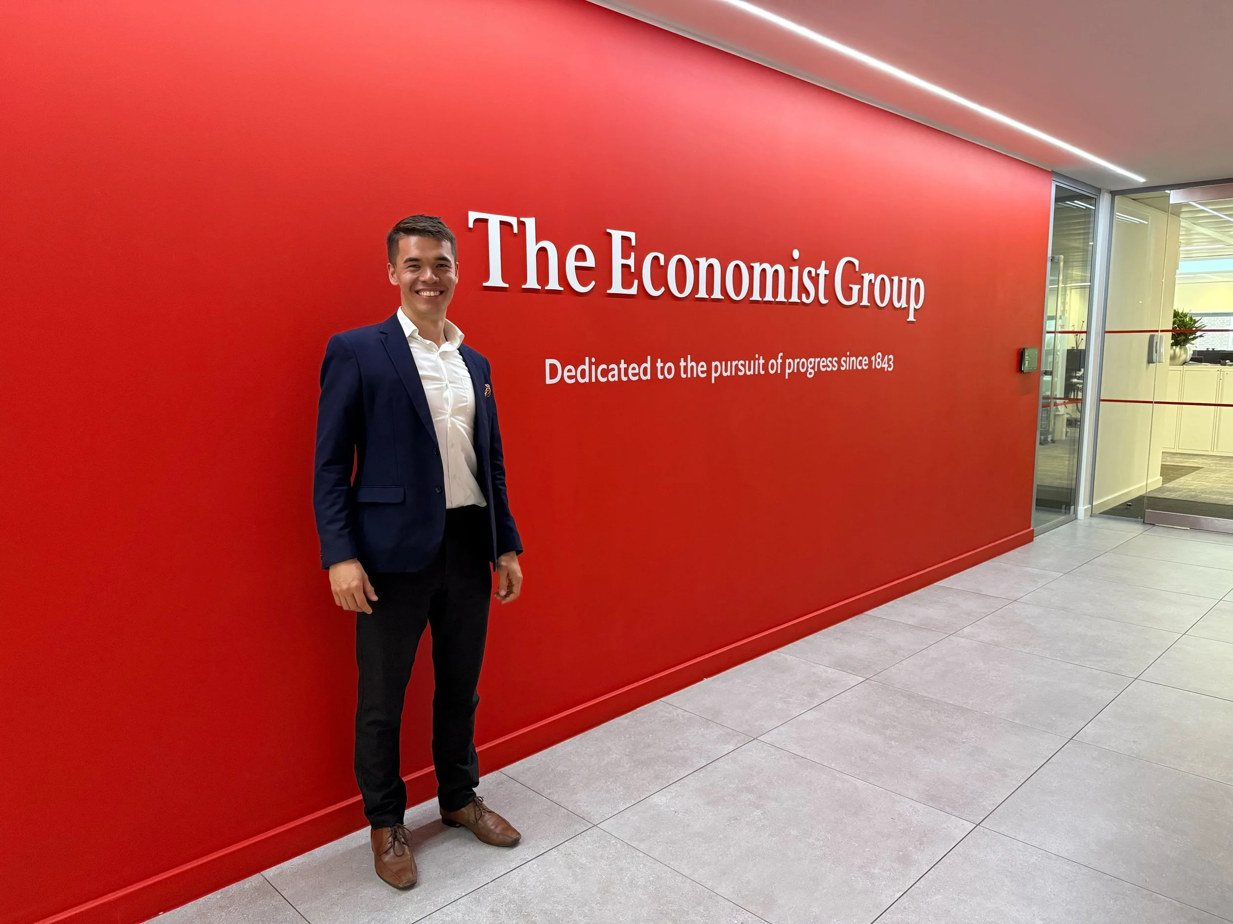 A man in a suit and white shirt standing in front of a red wall with white text that reads 'The Economist Group' and a motto 'Dedicated to the pursuit of progress since 1843' inside a modern office building.