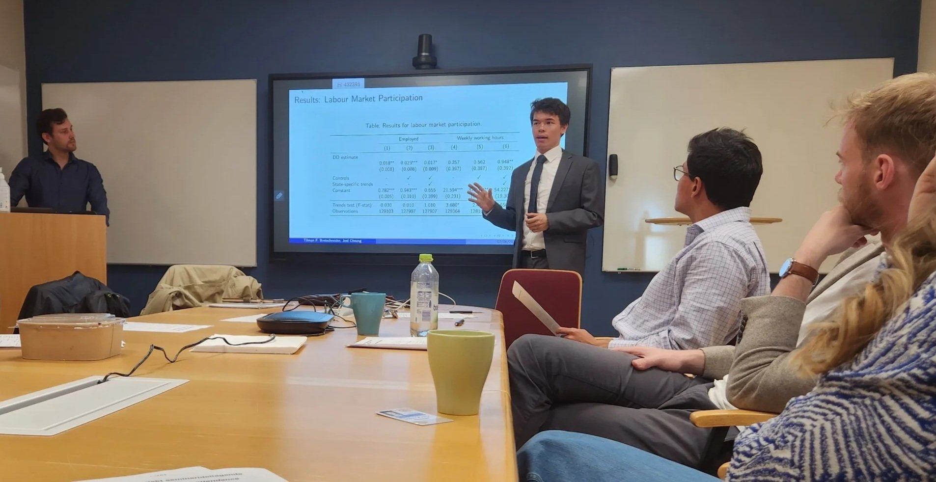A man in a gray suit and white shirt presenting in a conference room with a large screen displaying a PowerPoint slide on labor market participation. Several attendees are seated, listening to him, with notebooks, cups, and a water bottle on the table.