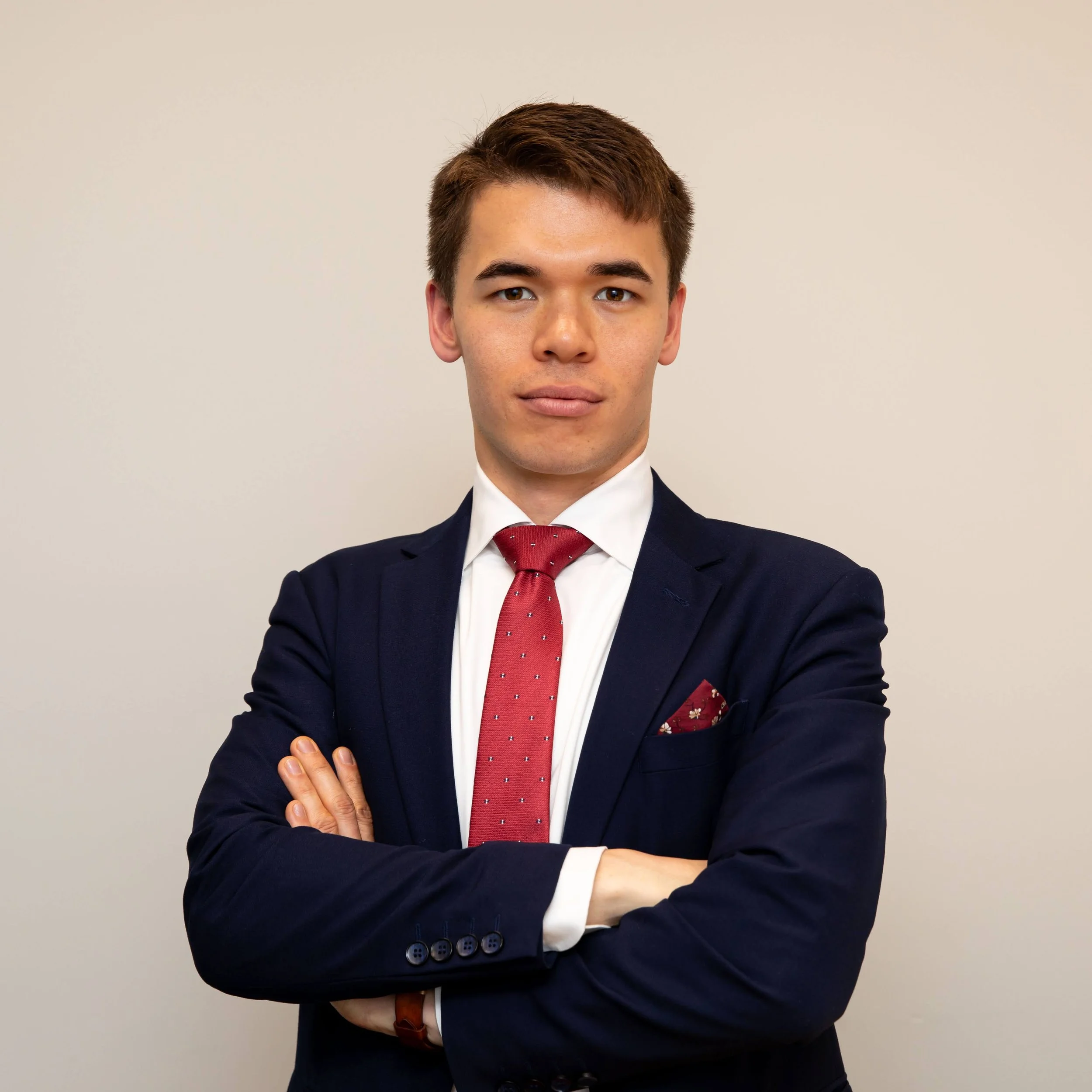 A young man wearing a dark suit with a red tie standing in front of a plain wall.